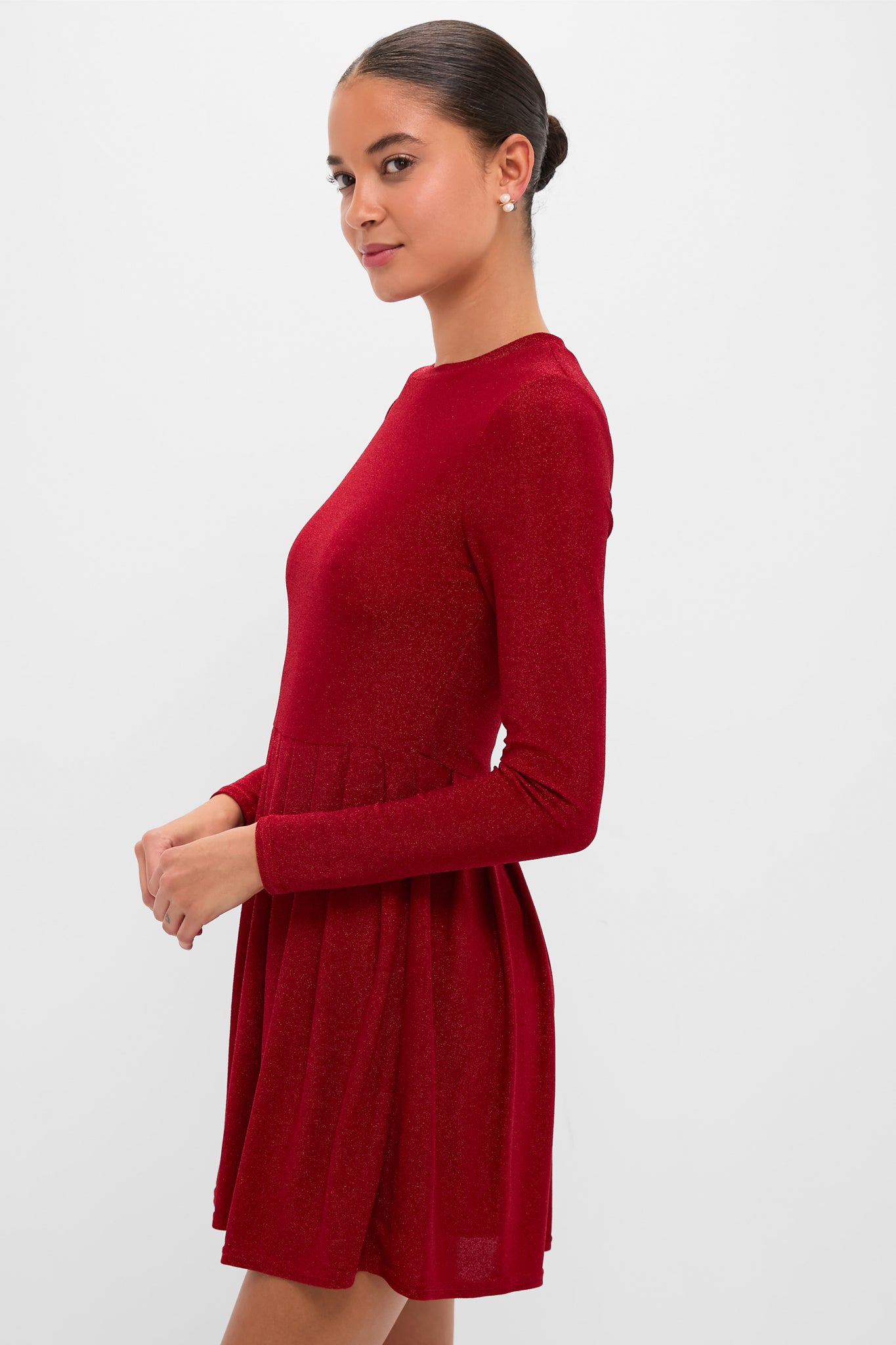 Woman models the Red Shimmer Pleated Bale Mini Dress, showcasing its classic, high-end design with an effortless, fun sparkle and elegant pleated skirt.