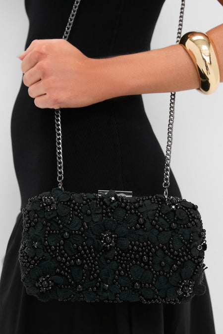 Woman in a classic black dress holding the Black Beth Encrusted Clutch, featuring effortless floral beadwork and a fun, high-end look with a gunmetal chain strap.