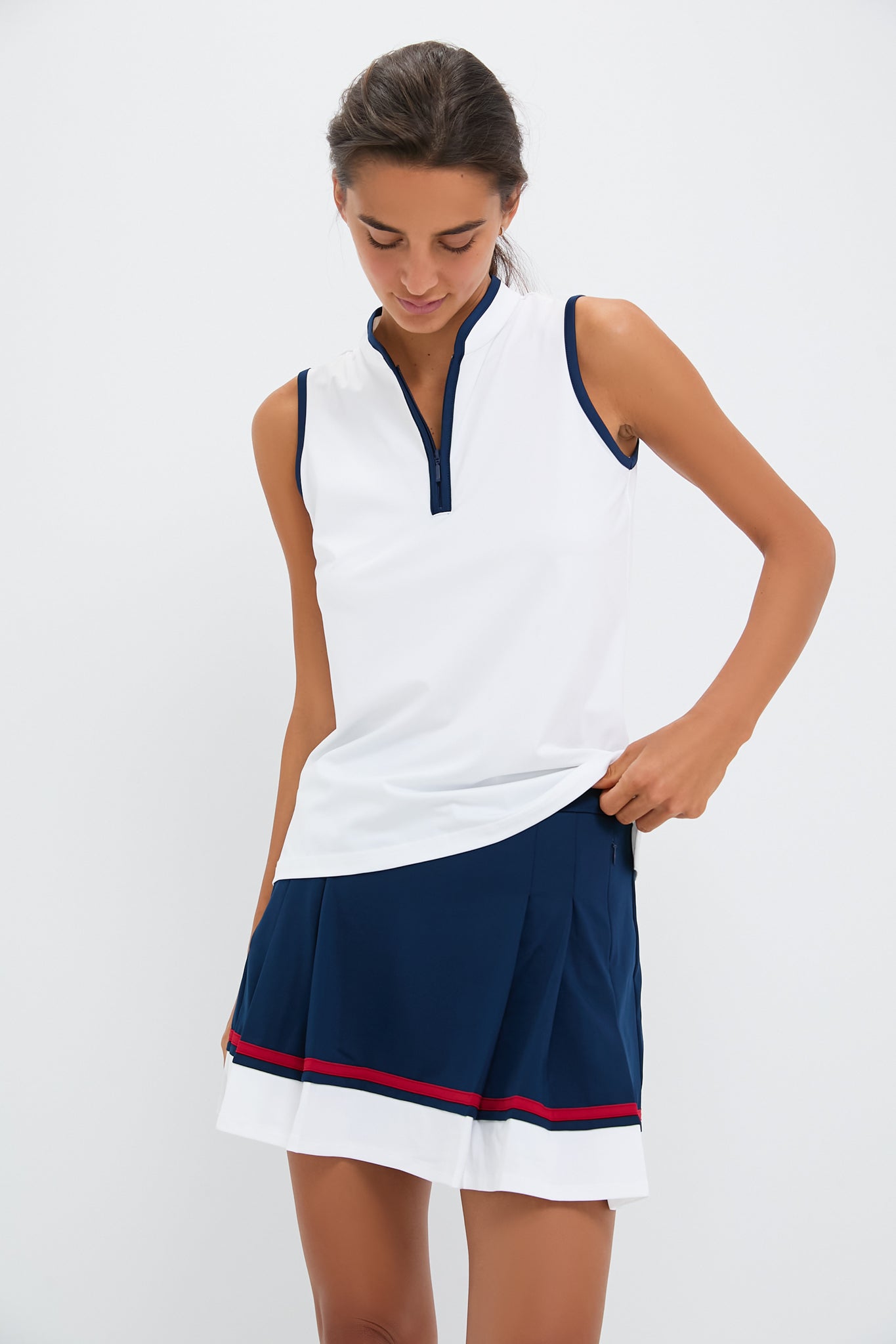 A woman wearing a Tnuck Sport sleeveless mock neck white top with a zippered collar and a navy pleated skirt with red and white stripes looks down, holding the hem of her top.