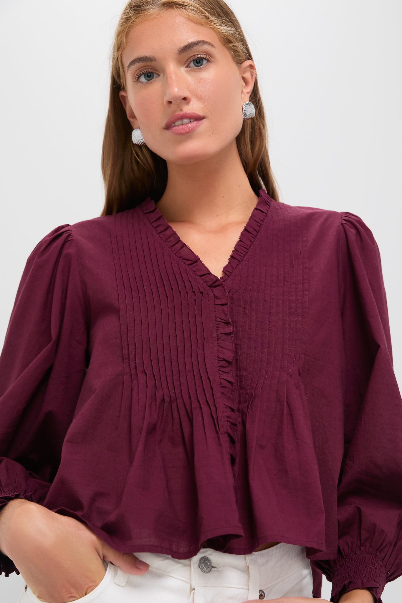 Young woman models the Exclusive Burgundy Anise Long Sleeve Top with pintuck pleats, puff shoulders, and smocked cuffs, paired effortlessly with light pants for a classic, high-end look.