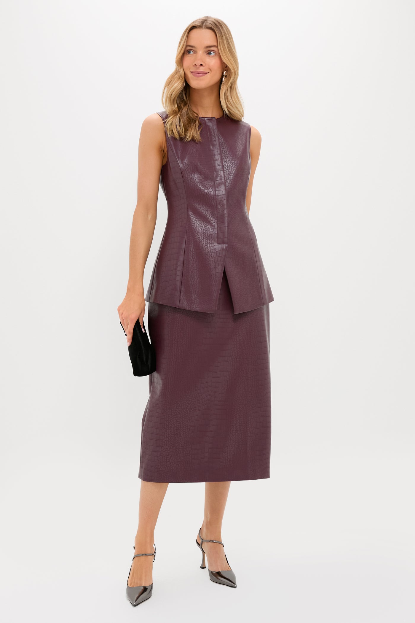 A woman poses against a plain background in a Tuckernuck sleeveless dark mauve textured top with a matching midi skirt, holding a black clutch and wearing silver pointed-toe heels.