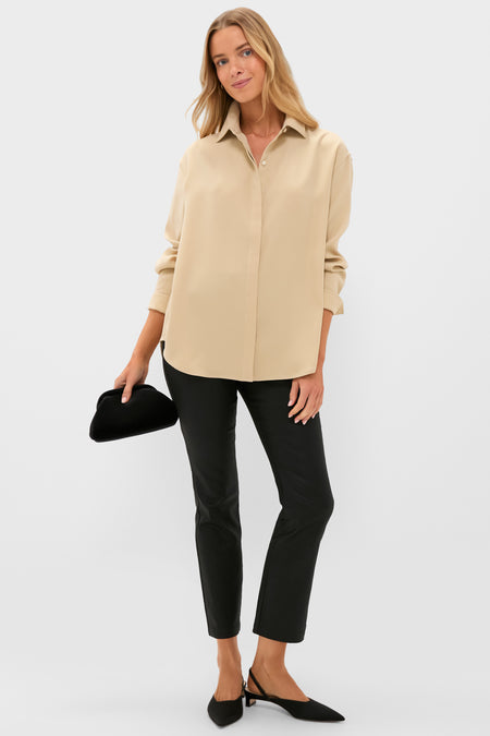 A woman stands against a plain background wearing a Tuckernuck relaxed fit beige blouse, black pants, black slingback shoes, and holding a black hat.