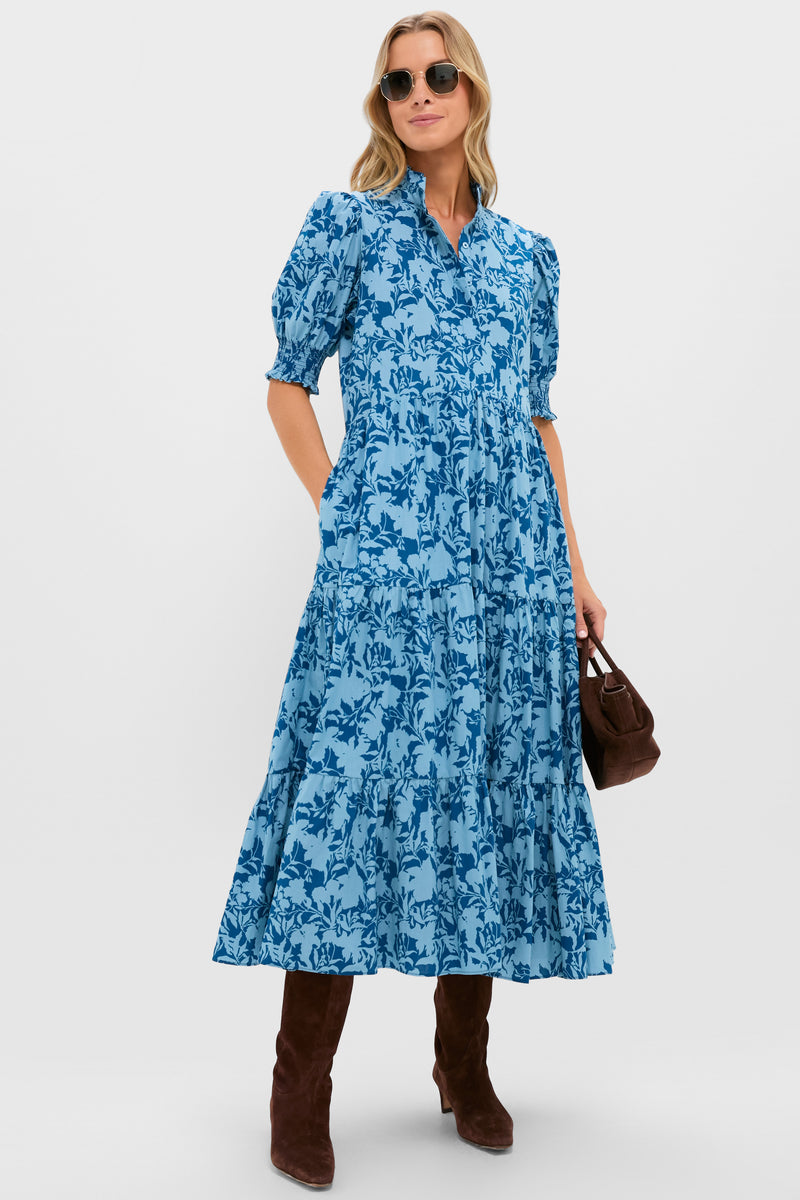 A woman in a steel blue floral maxi dress by Hyacinth House, featuring a tiered midi skirt and ruffled stand collar, wears sunglasses, brown knee-high boots, and carries a brown bag against a plain background.