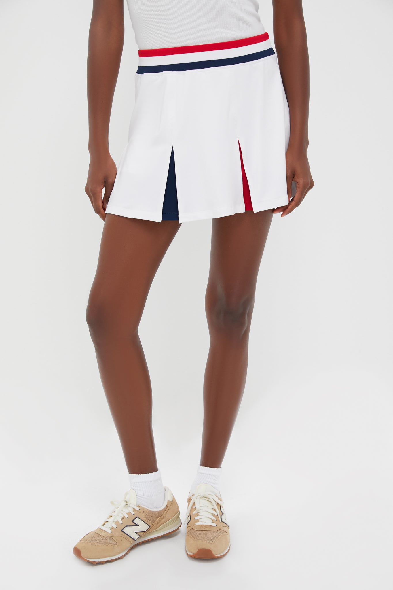 A person in a Tnuck Sport white tennis skirt with navy and red accents, paired with a white top, white socks, and beige New Balance sneakers showcases quick-dry comfort and athletic style with confidence.