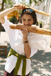 Woman holding two baguettes in a white button down and burgundy pants with a green sweater tied around her waist. 