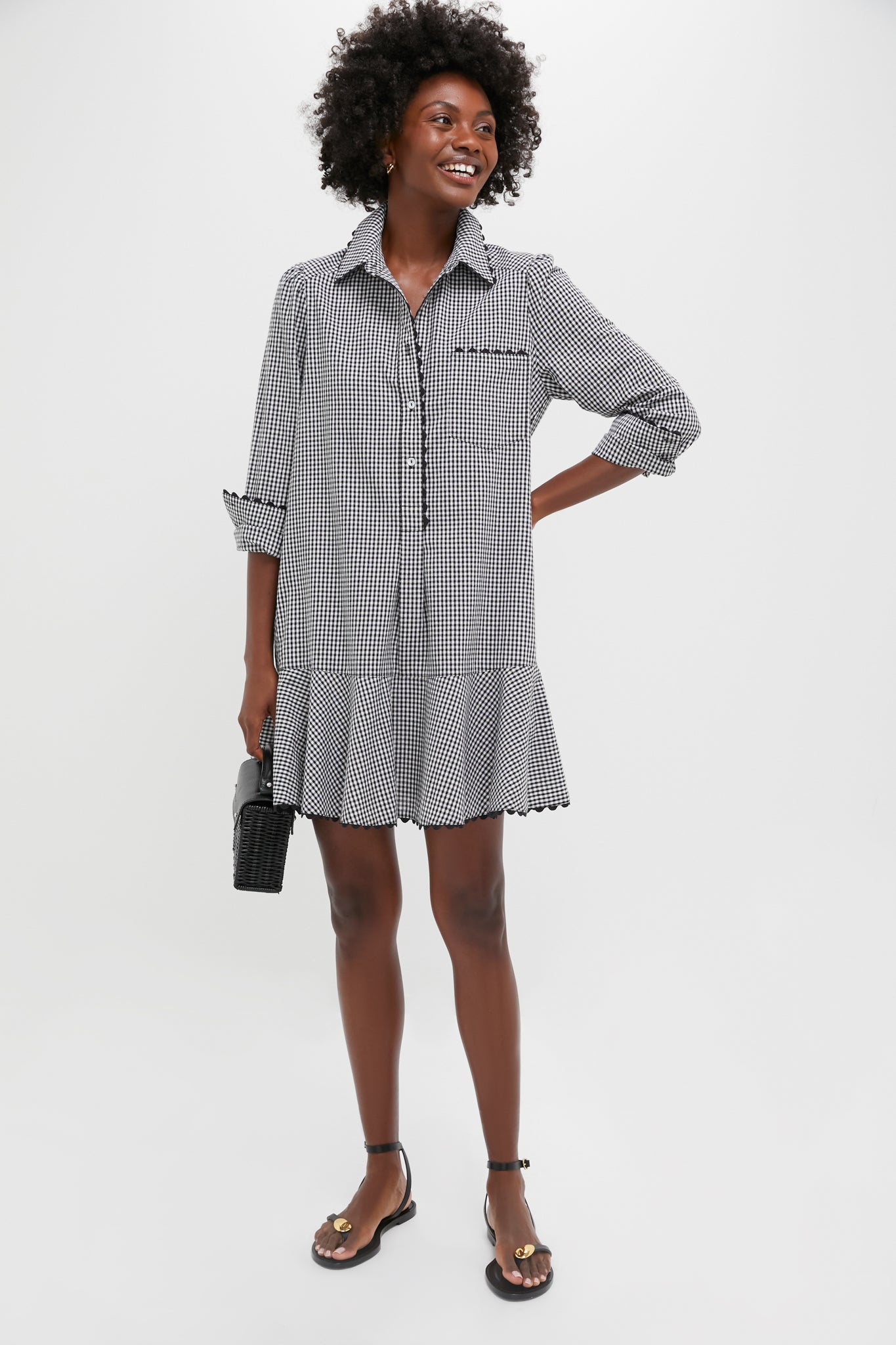 A woman wears the Tuckernuck Black Gingham Callahan Shirt Dress with a relaxed fit, collared neckline, long sleeves, ruffled hem, black sandals, and carries a small black handbag.