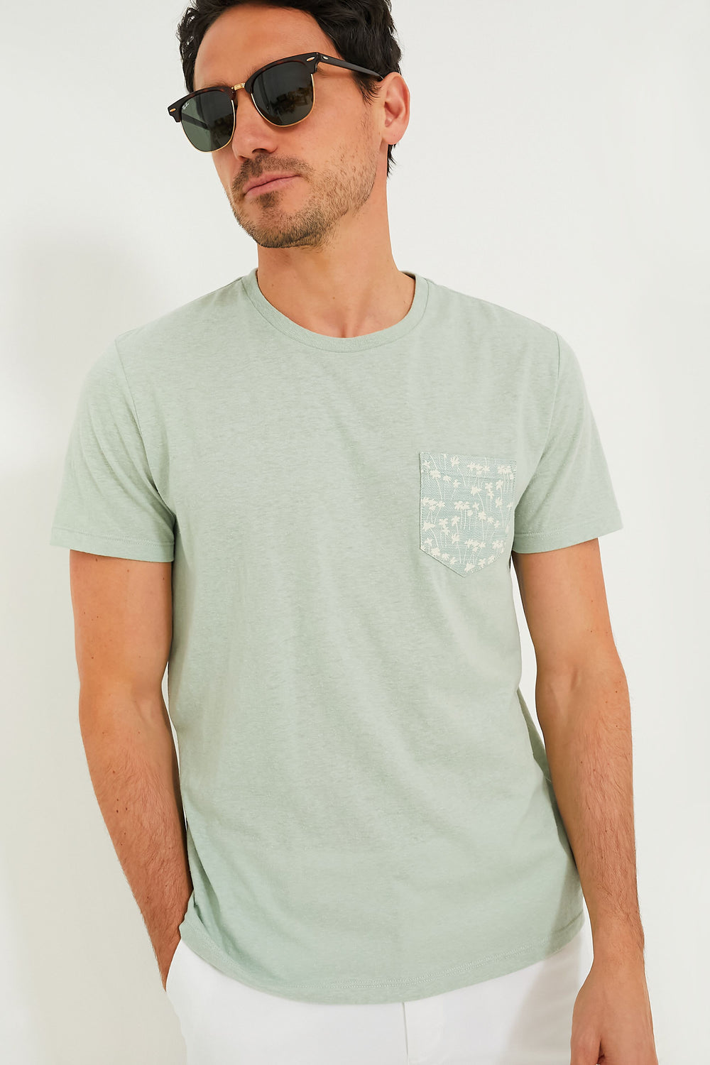 A man stands confidently wearing black sunglasses, white pants, and a light green Marine Layer eco-friendly t-shirt featuring a patterned chest pocket.