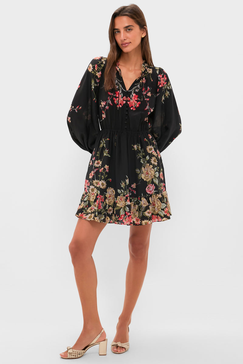 A woman stands with one hand on her hip, wearing a black floral mini dress with long sleeves, a cinched waist and hem from Sea New York, paired with beige heeled sandals.