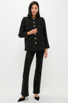 A woman stands against a white background wearing a black tweed blazer with gold buttons, black pants, and black heels by Pomander Place.