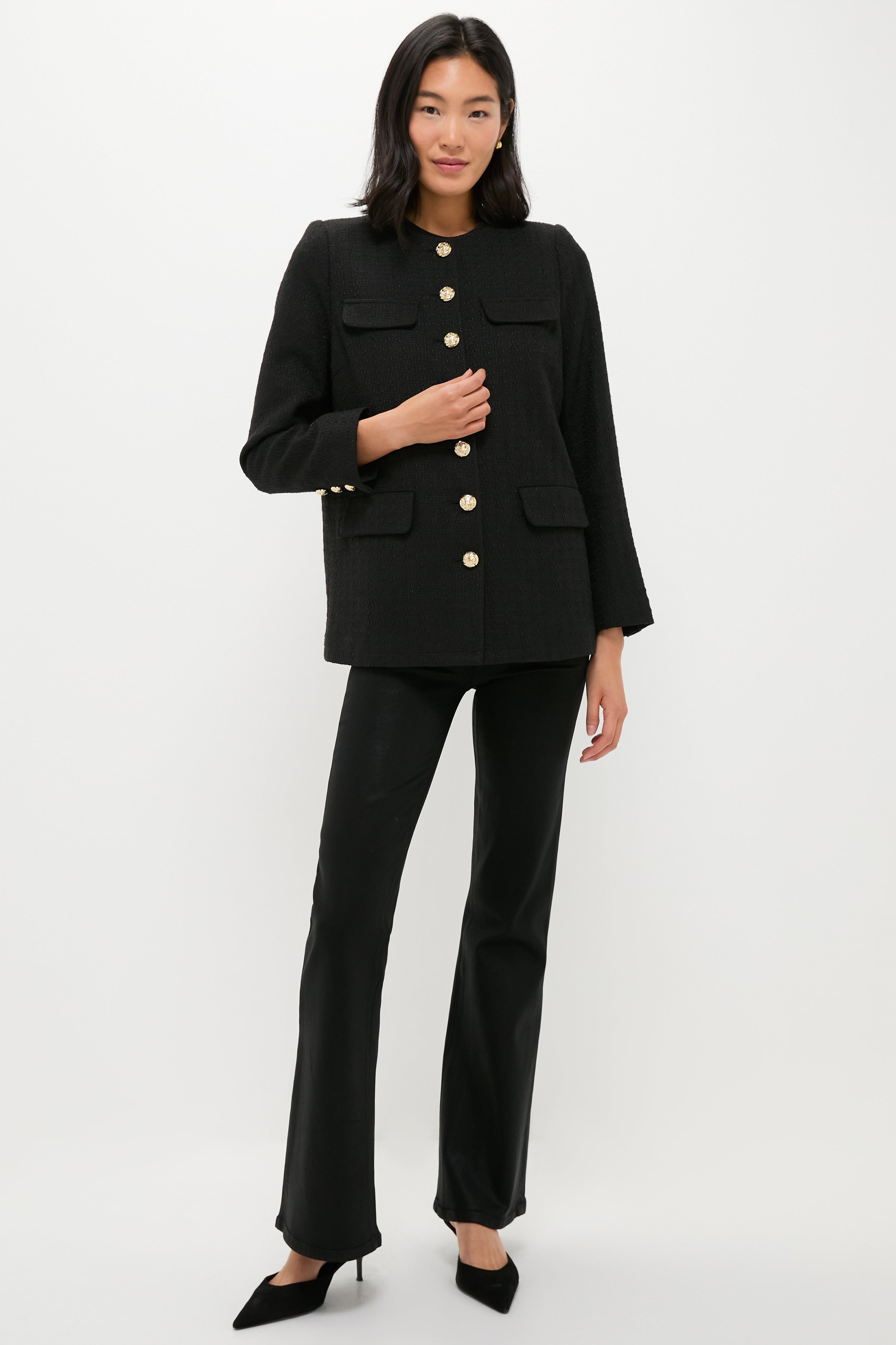 A woman stands against a white background wearing a black tweed blazer with gold buttons, black pants, and black heels by Pomander Place.