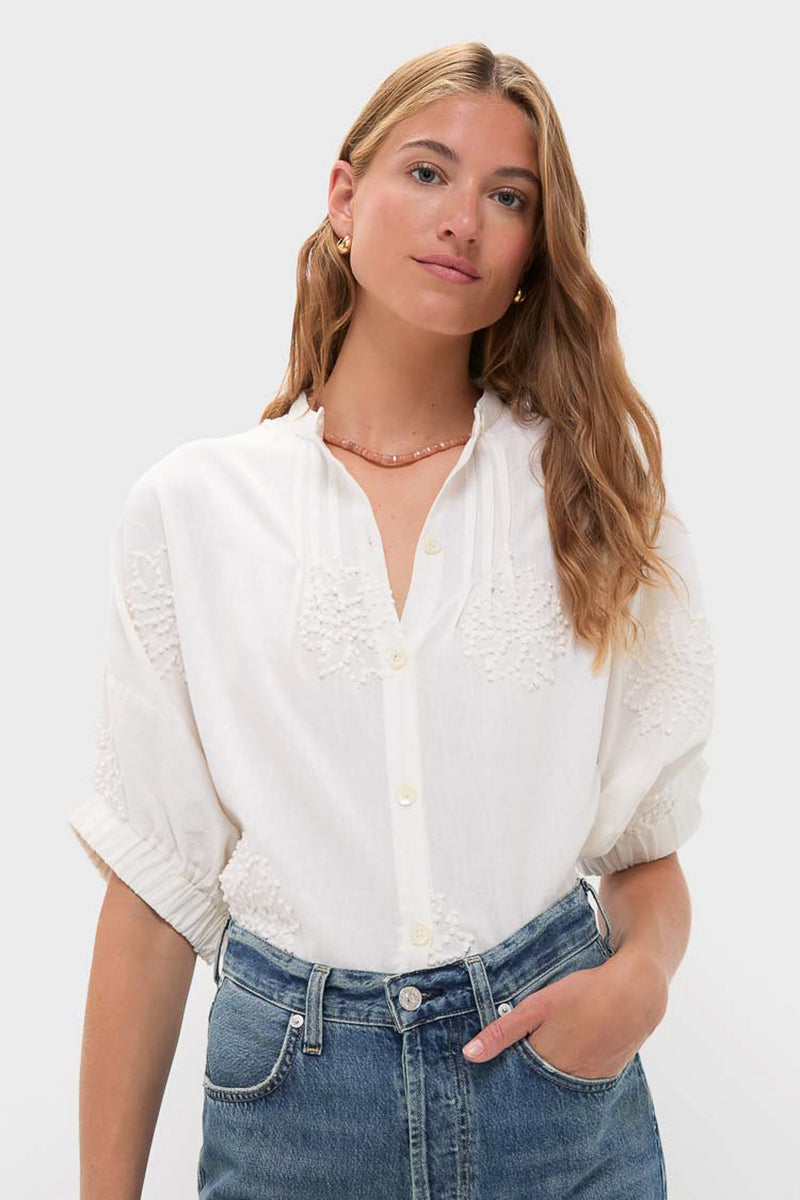 A woman with light brown hair wears a hand-stitched cotton flax blouse with embroidered details by Love Tomas by Emerson Fry and blue jeans, standing against a plain light gray background.