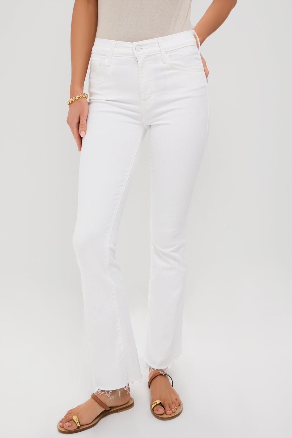Wearing high-waisted, white MOTHER Weekender Fray jeans with frayed hems, a beige top, gold bracelet, and tan sandals for an effortlessly stylish look.