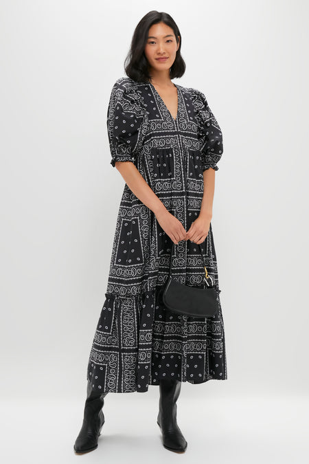 A woman stands against a plain background wearing a Hunter Bell black and white patterned maxi dress, black boots, and holding a black handbag.