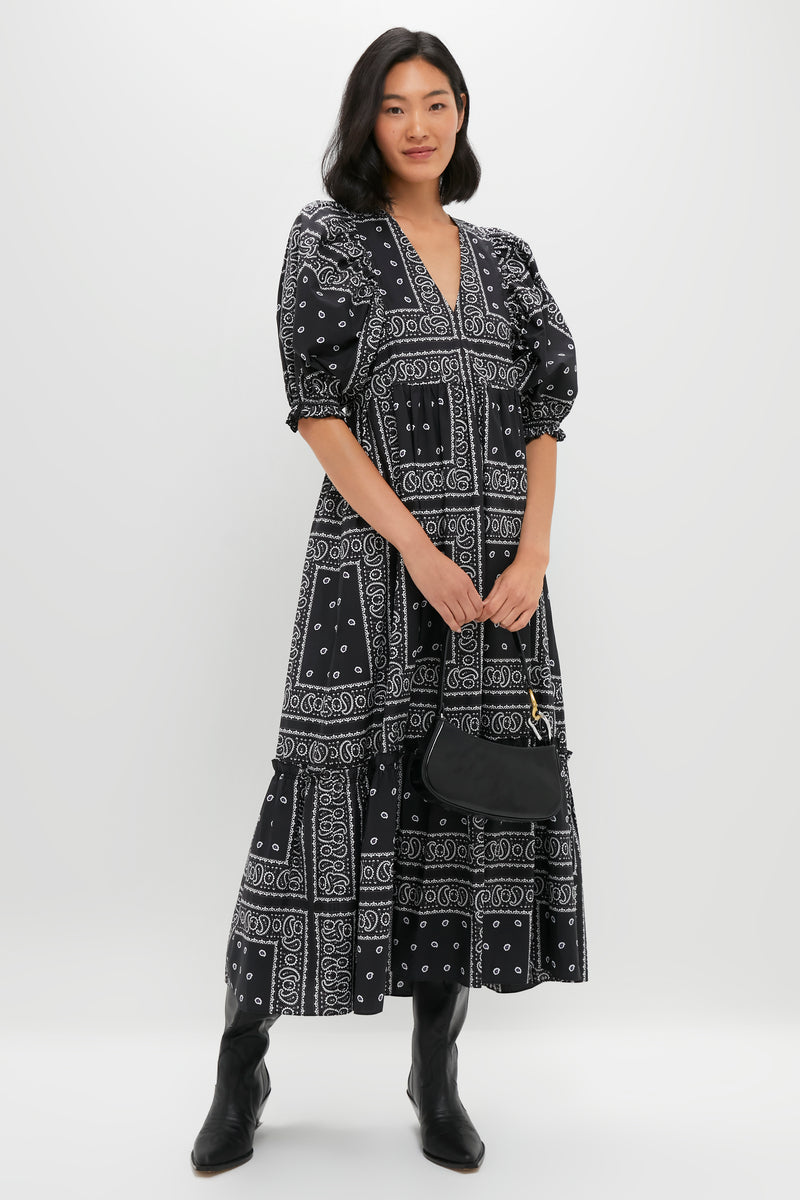 A woman stands against a plain background wearing a Hunter Bell black and white patterned maxi dress, black boots, and holding a black handbag.