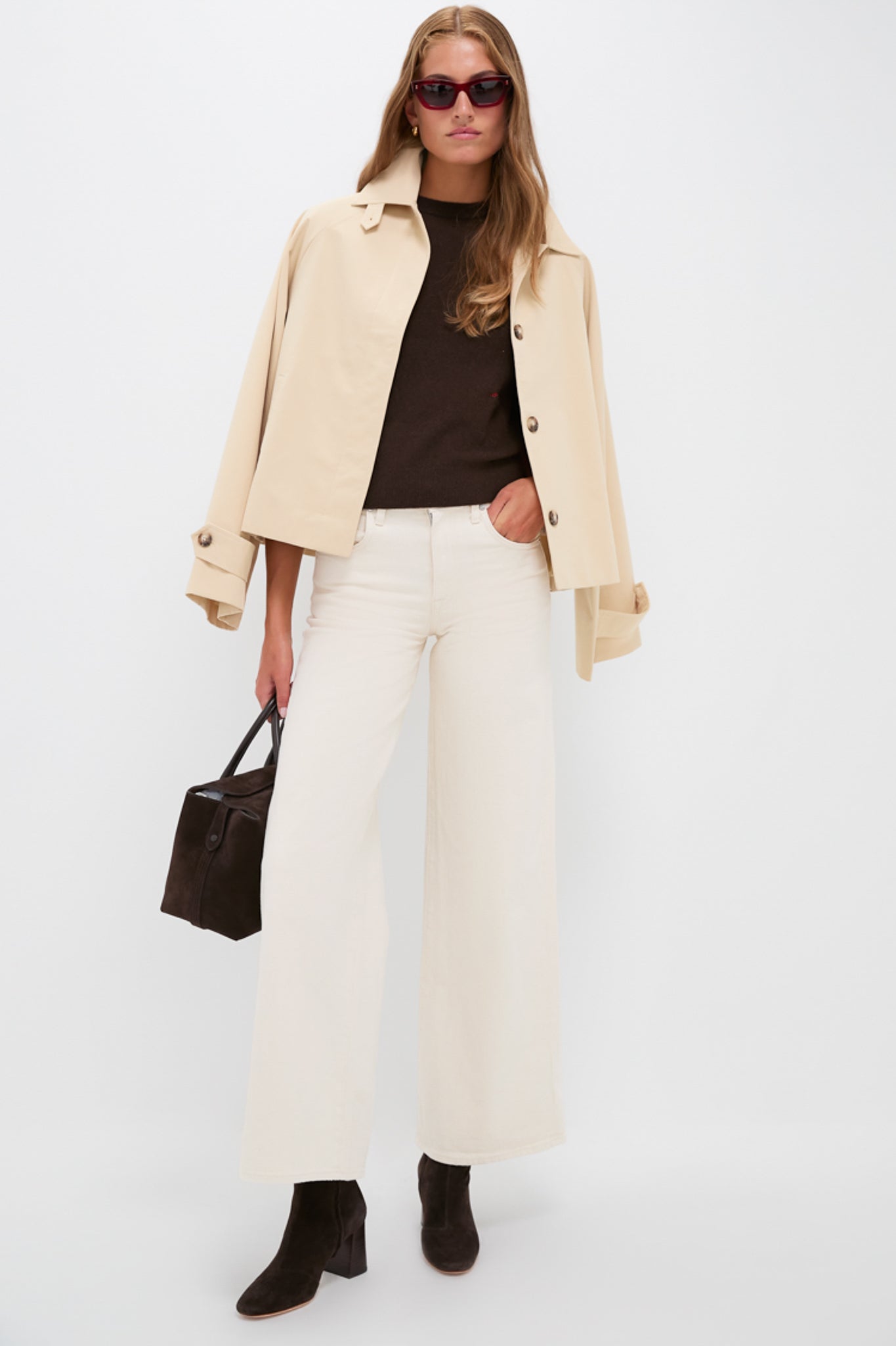 A woman in a Tuckernuck beige jacket, chocolate brown cashmere crewneck, white wide-leg pants, brown boots, sunglasses, and holding a dark bag stands against a plain white background.
