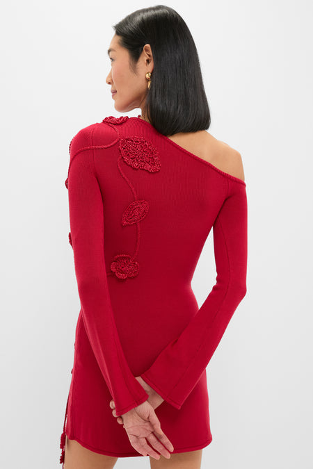 A woman poses with hands behind her back against a plain background, wearing a red, one-shoulder Cult Gaia dress featuring long sleeves and 3D floral applique detailing on the back.