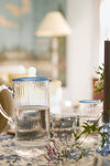 Chambray Nell Pitcher, a classic, high-end glass piece with a blue rim, effortlessly elevates any table setting, surrounded by matching glasses and a floral tablecloth.