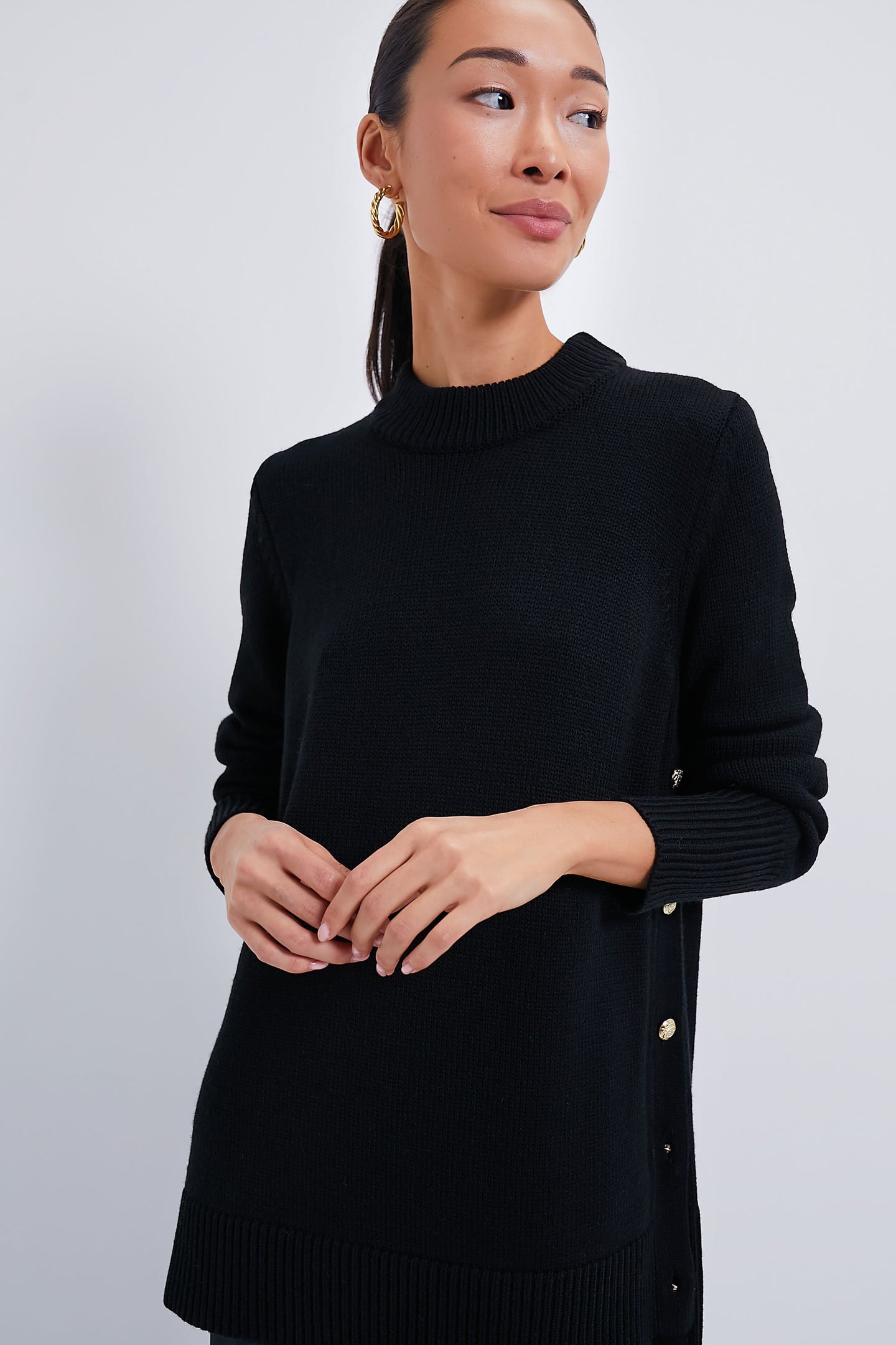 A woman wears the Tuckernuck Black Bexley Sweater with gold side buttons and a high-low hem, paired with gold hoop earrings.