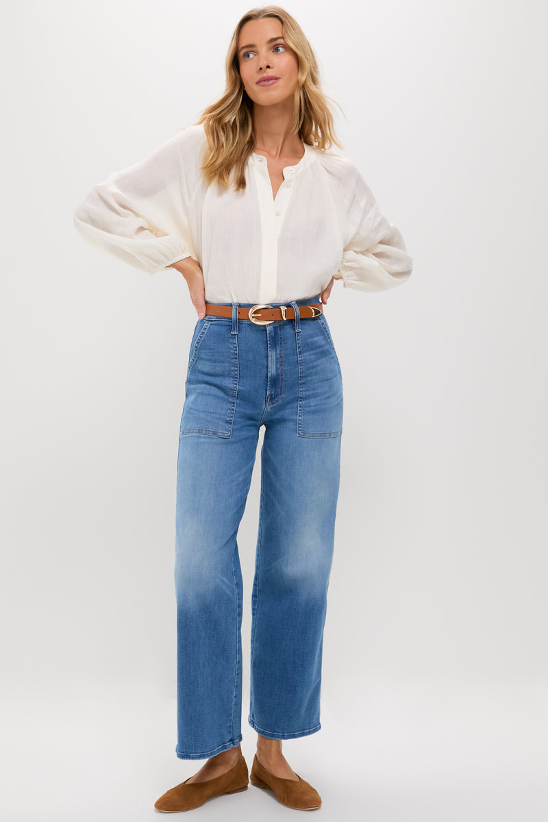 A woman stands against a plain background wearing a white blouse, MOTHER high-rise wide leg jeans in recycled stretch denim with front pockets, a brown belt, and brown flats.