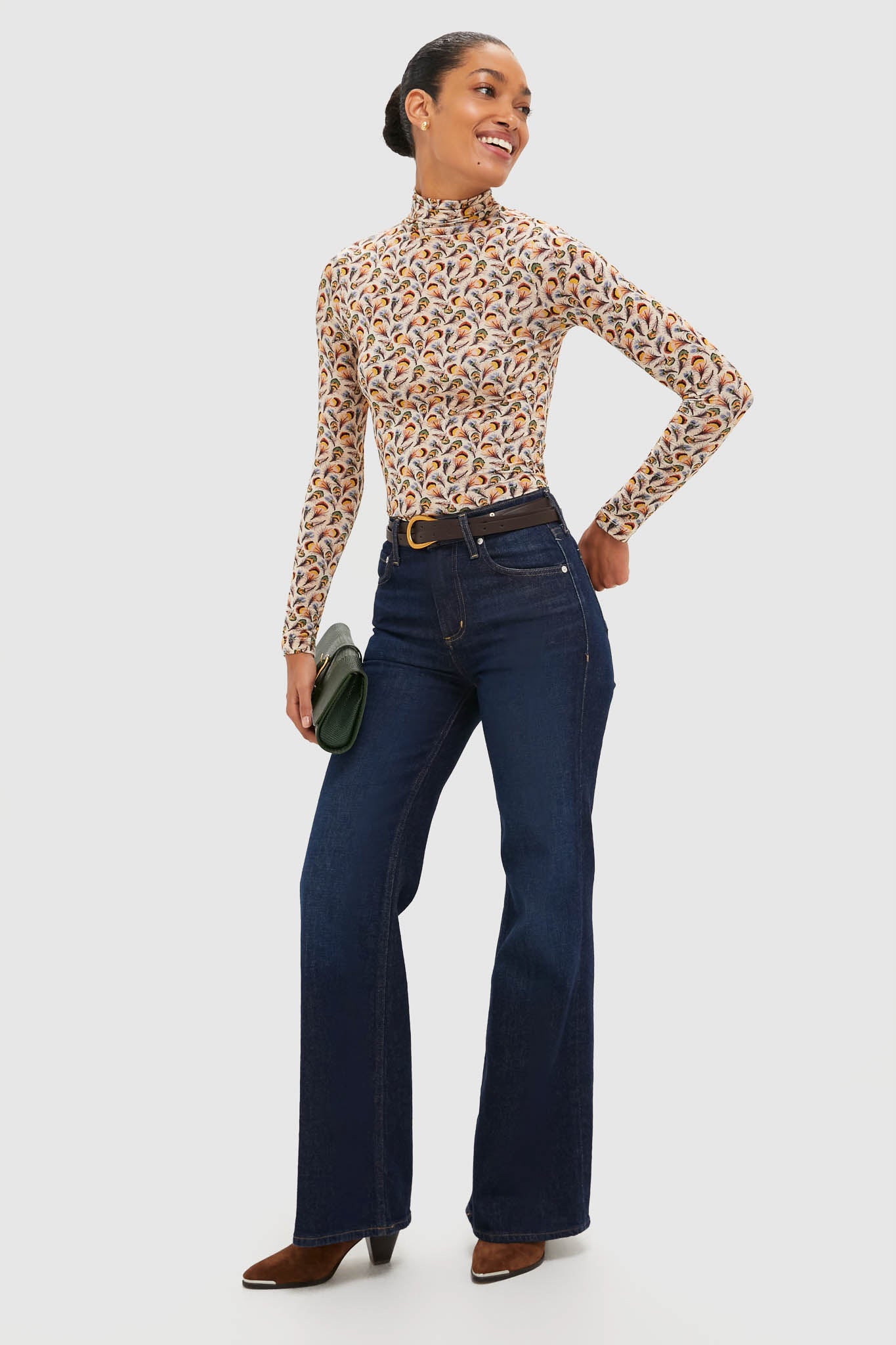 A woman smiles against a plain background, wearing the Ulla Johnson Aurelia Turtleneck in lightweight tricot jersey with dark high-waisted flare jeans, brown boots, and holding a green clutch.