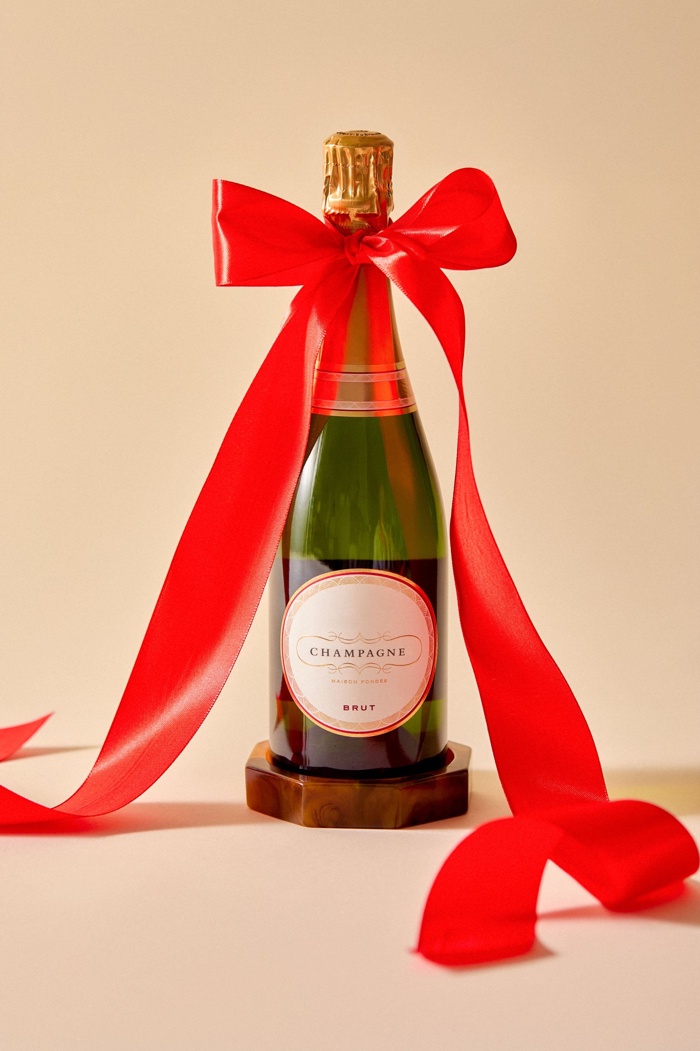 A Tuckernuck Home champagne bottle with a gold foil top and large red ribbon stands upright on a beige surface beside elegant resin tortoise shell wine coasters.