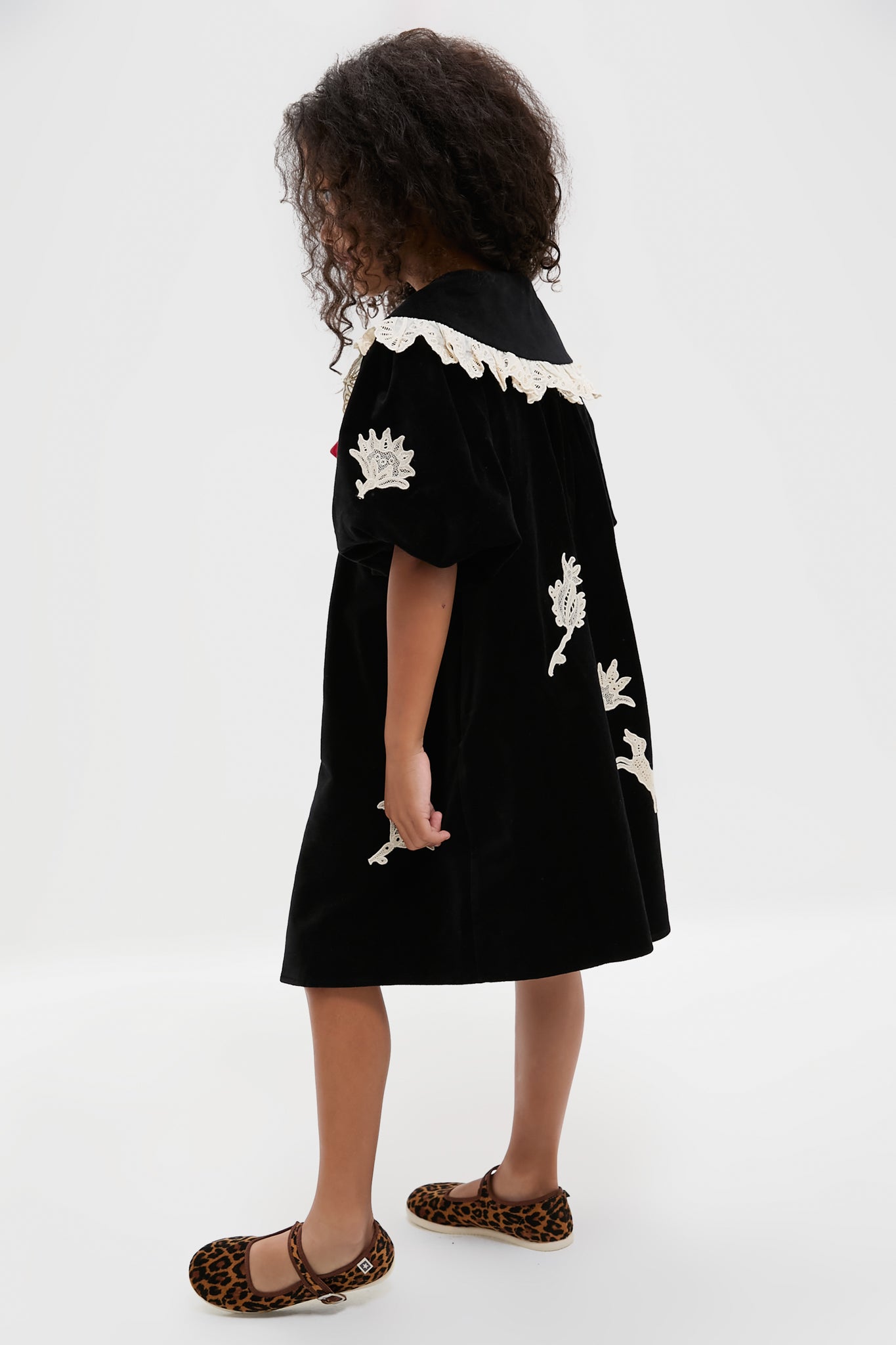 A young girl stands sideways in a Sea New York velveteen lace applique dress with white embroidered details, paired with leopard print flat shoes.