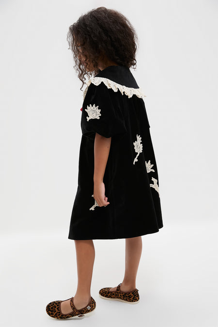 A young girl stands sideways in a Sea New York velveteen lace applique dress with white embroidered details, paired with leopard print flat shoes.
