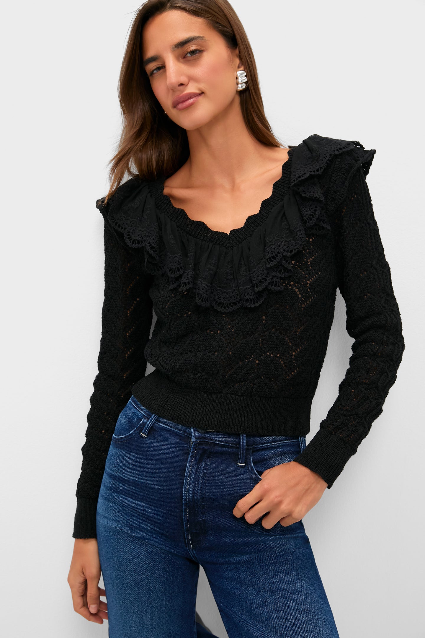A woman stands confidently in a Hyacinth House black ruffled V-neck sweater with lace details, paired with blue jeans.