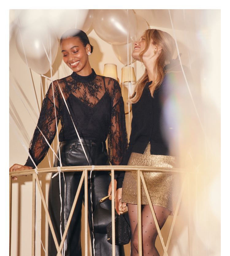 An image of two women standing on a balcony. The woman on the left wears a black sheer lace top and leather pants, styled with a black top handle purse. The woman on the right wears a black cashmere cardigan, a gold jacquard mini skirt, and polka dot tights.