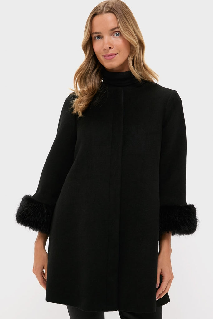 A woman stands against a plain light background, wearing a Tuckernuck Black Wool Coat with three-quarter sleeves and faux fur cuffs, adding elegance to her look.