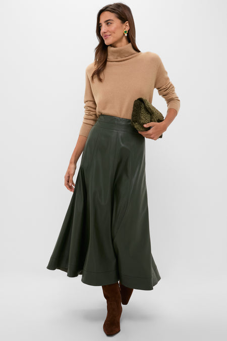A woman stands against a plain backdrop wearing a Tuckernuck cashmere turtleneck sweater, olive green A-line leather skirt, brown suede boots, and holding a green clutch.