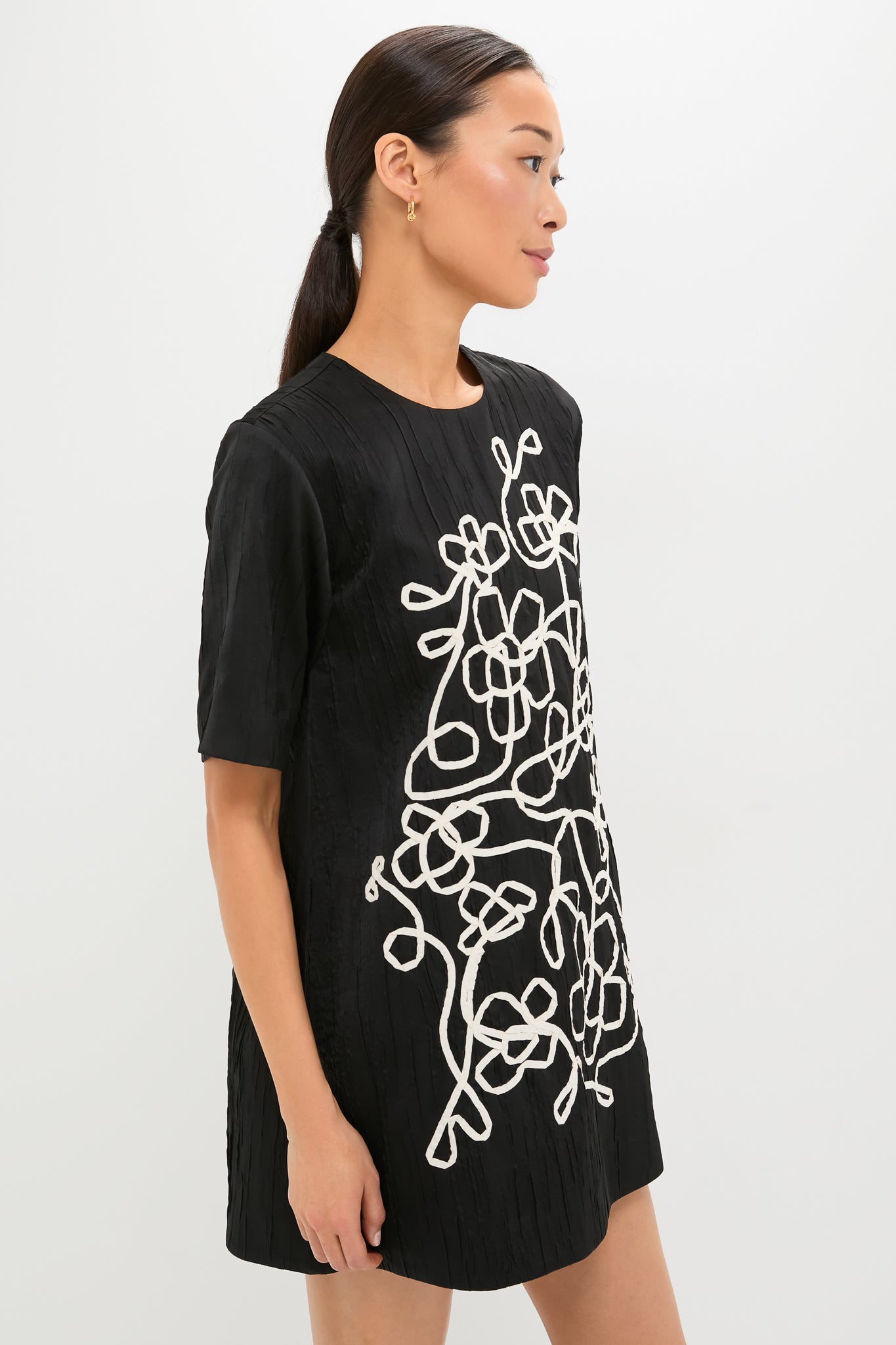 Woman models the Onyx and Ivory Classic Katya Mini Dress featuring effortless elegance with its textured fabric and abstract white vine embroidery, exuding a fun and high-end style.