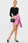 Model wearing the Pink Metallic Plisse Brenna Bow Mini Skirt with a fitted black sweater, paired with an effortless black sequin shoulder bag and classic slingback pumps.