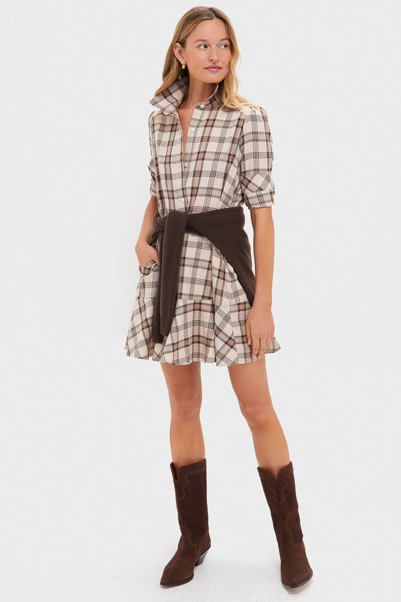 A woman wears a best-selling Tuckernuck plaid day dress with a dark sweater tied at the waist and brown knee-high boots, standing against a plain white background.