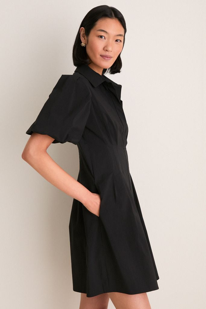 Woman in classic Black Mini Delaney Dress with puff sleeves and pleated waist, embodying effortless, high-end style for versatile fun occasions, hands in side seam pockets.