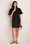 Woman models the Black Mini Delaney Dress, a classic, high-end shirt dress with puff sleeves and pleated waist, styled with black sandals and a woven handbag for an effortless, fun look.