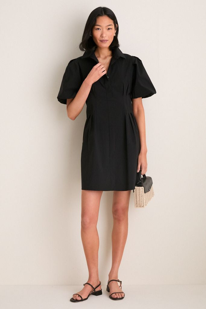 Woman models the Black Mini Delaney Dress, a classic, high-end shirt dress with puff sleeves and pleated waist, styled with black sandals and a woven handbag for an effortless, fun look.