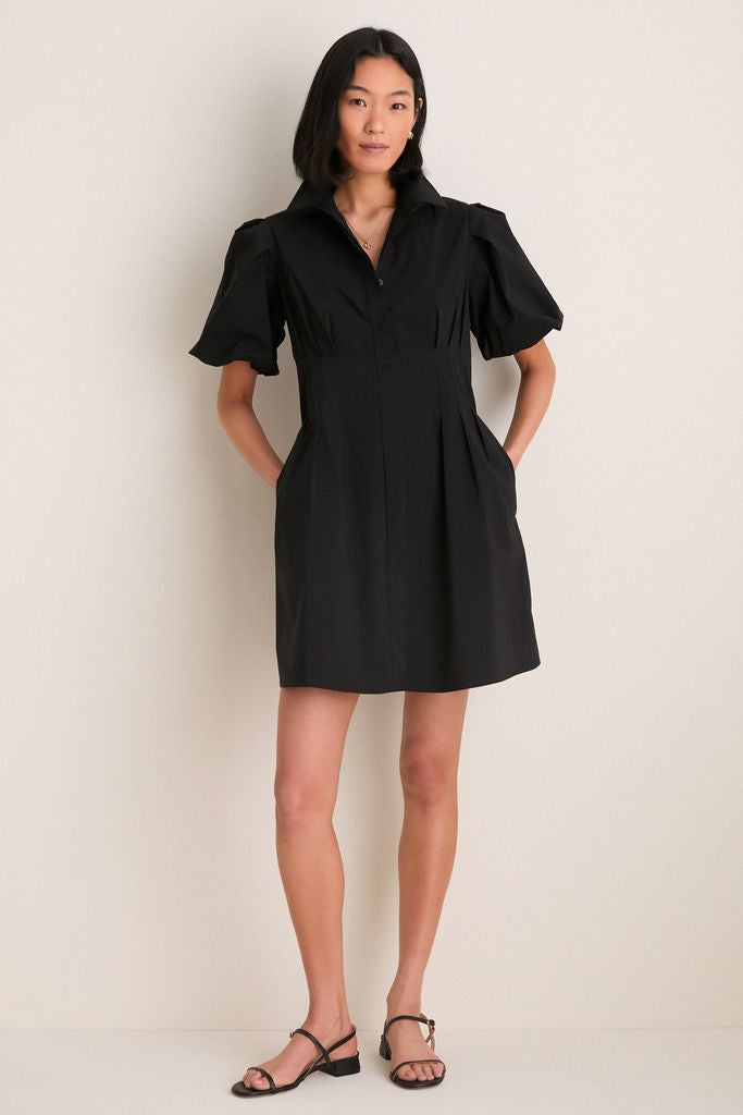 Woman in Black Mini Delaney Dress with puff sleeves and pleated waist, paired with strappy sandals, showcasing an effortless, classic look for versatile, high-end style.