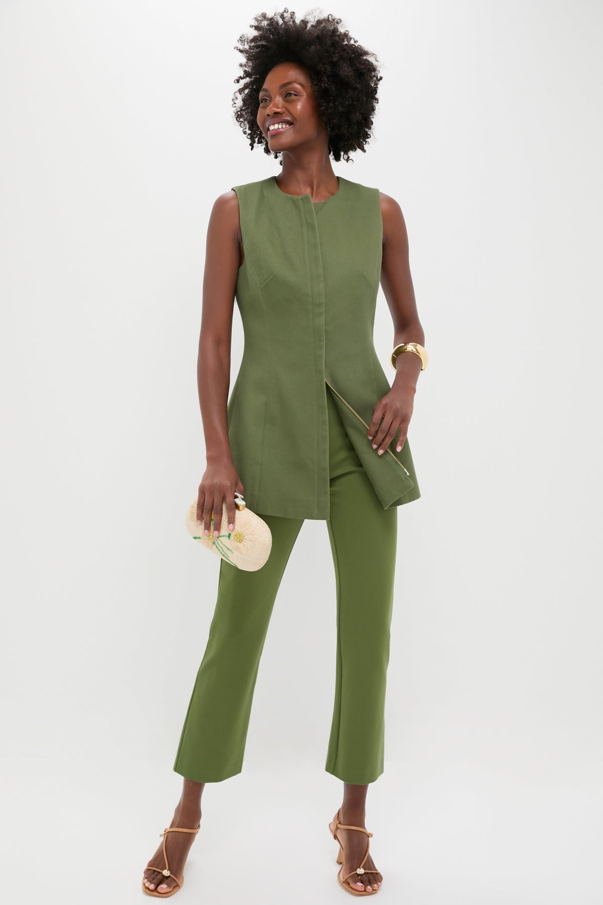 A woman dons the Tuckernuck Green Rhoda Tunic Top with matching pants, paired with a beige clutch and strappy heels, showcasing a chic, classic look.