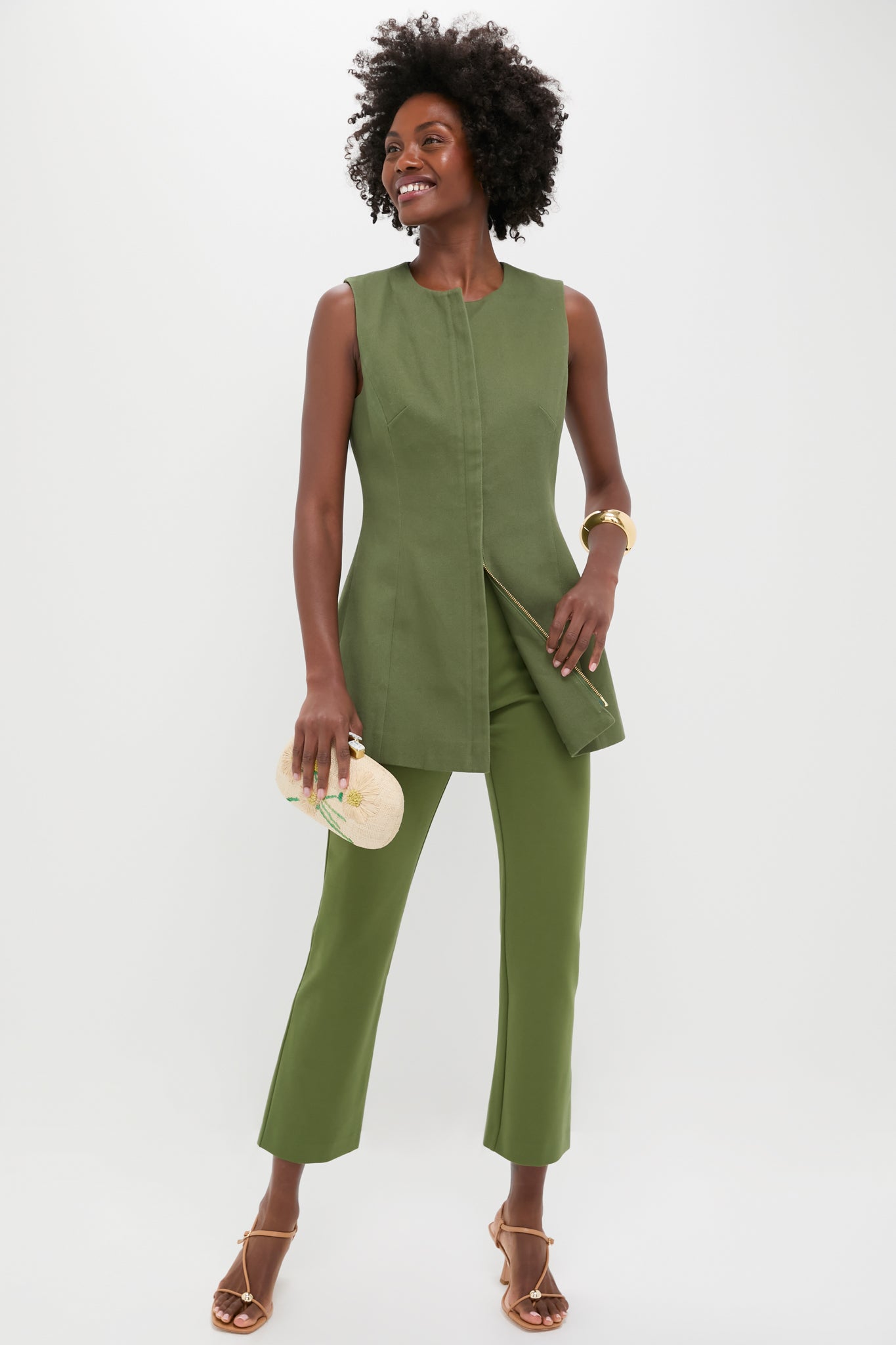 A woman dons the Tuckernuck Green Rhoda Tunic Top with matching pants, paired with a beige clutch and strappy heels, showcasing a chic, classic look.