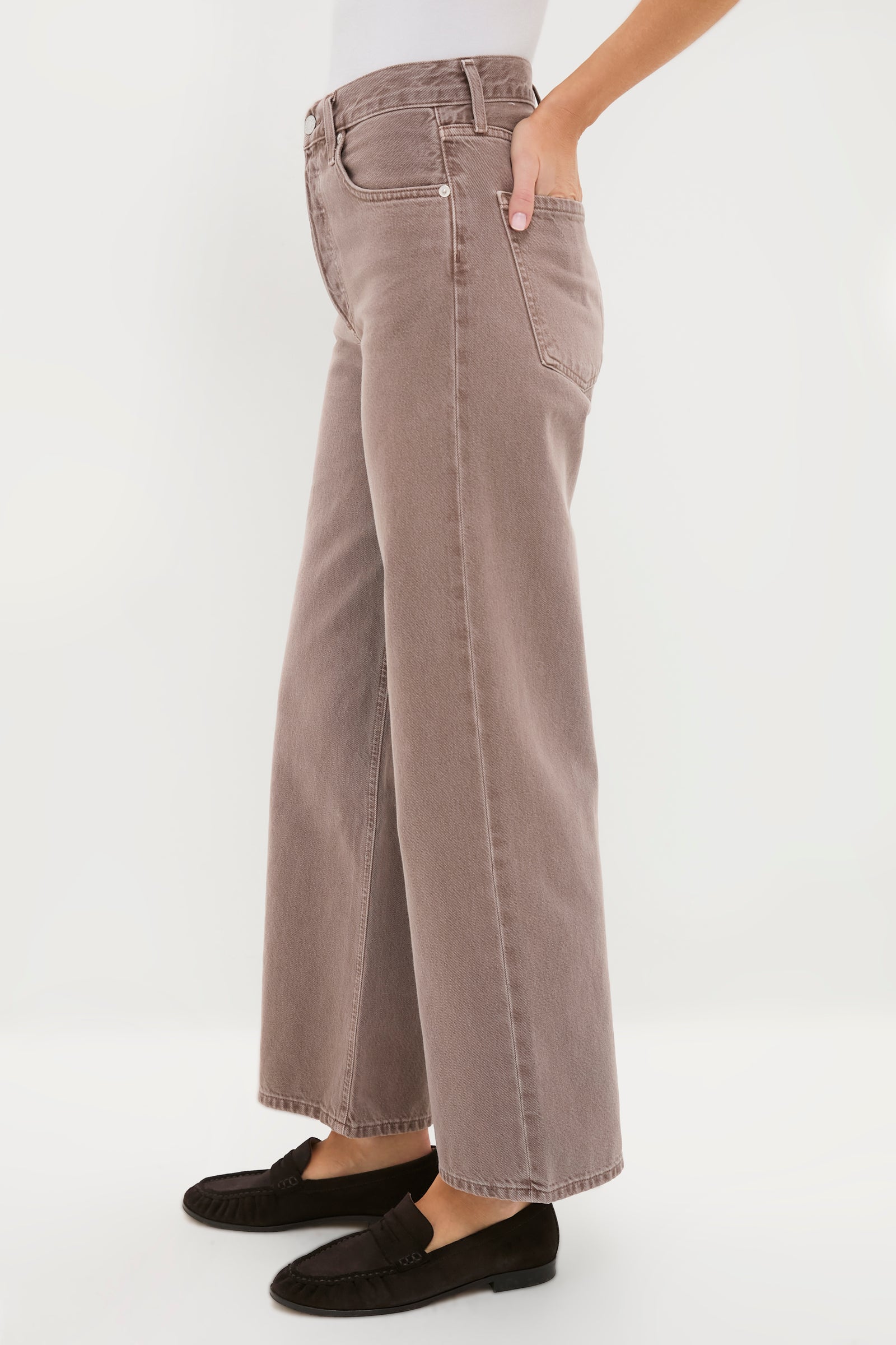 A person wears AGOLDE wide-leg, high-rise mauve jeans made from regenerative cotton with black loafers, hand in pocket, against a plain white background.