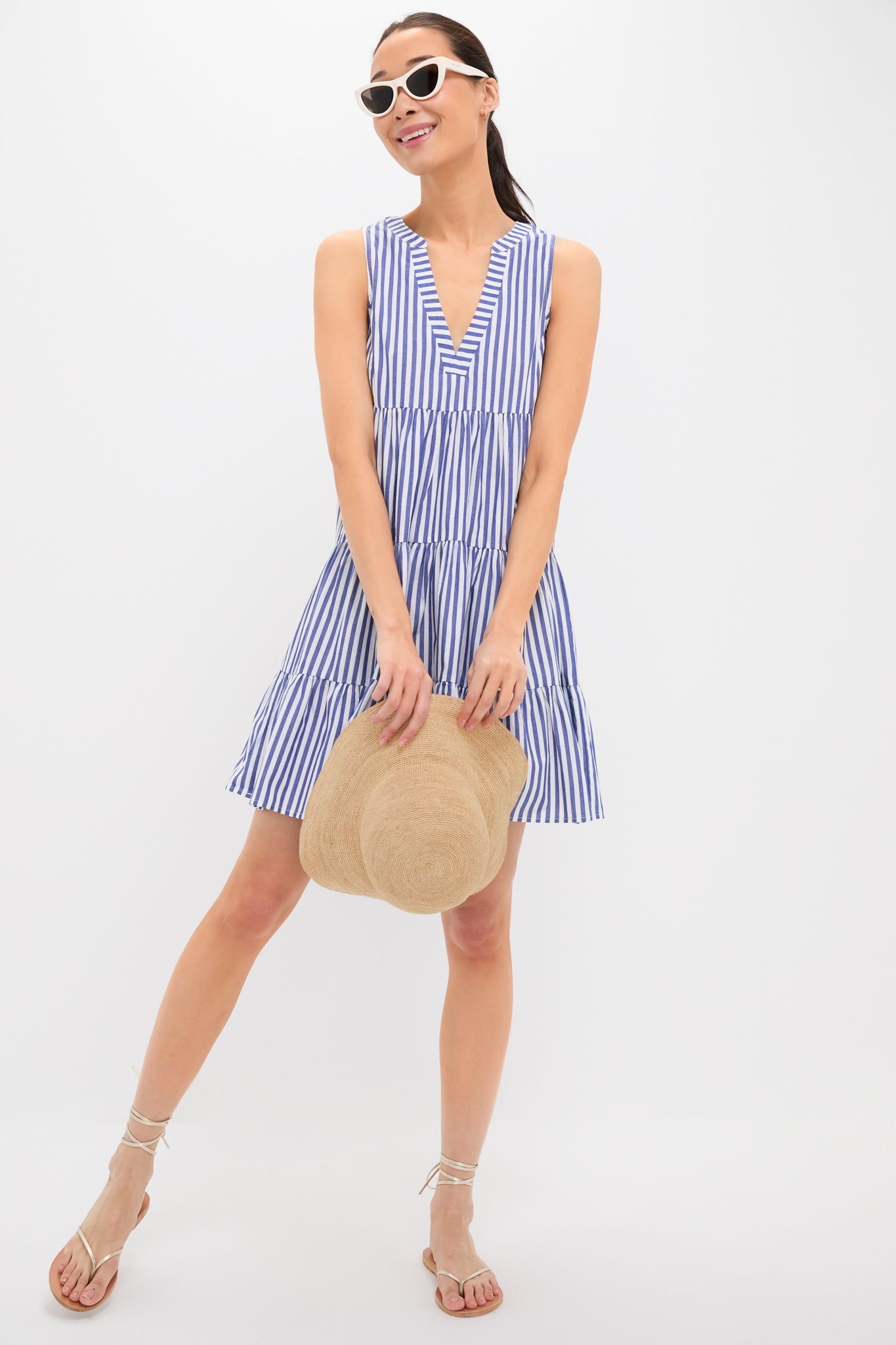 Woman wearing a sleeveless blue and white cotton poplin Pomander Place dress with stripes, white sunglasses, and sandals, holding a straw hat.