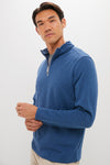 A man stands against a plain background in a blue Peter Millar quarter-zip pullover, layered over a gray shirt and beige pants, looking slightly to the side.