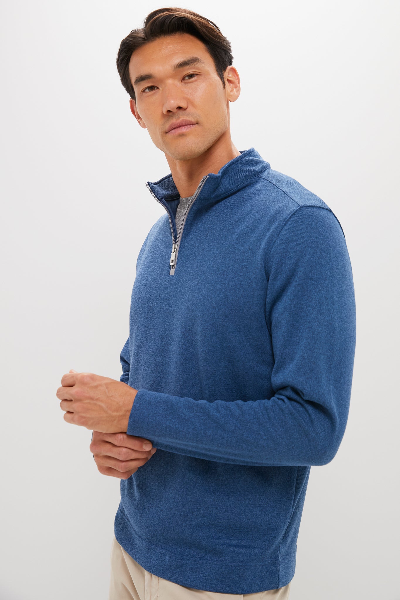 A man stands against a plain background in a blue Peter Millar quarter-zip pullover, layered over a gray shirt and beige pants, looking slightly to the side.