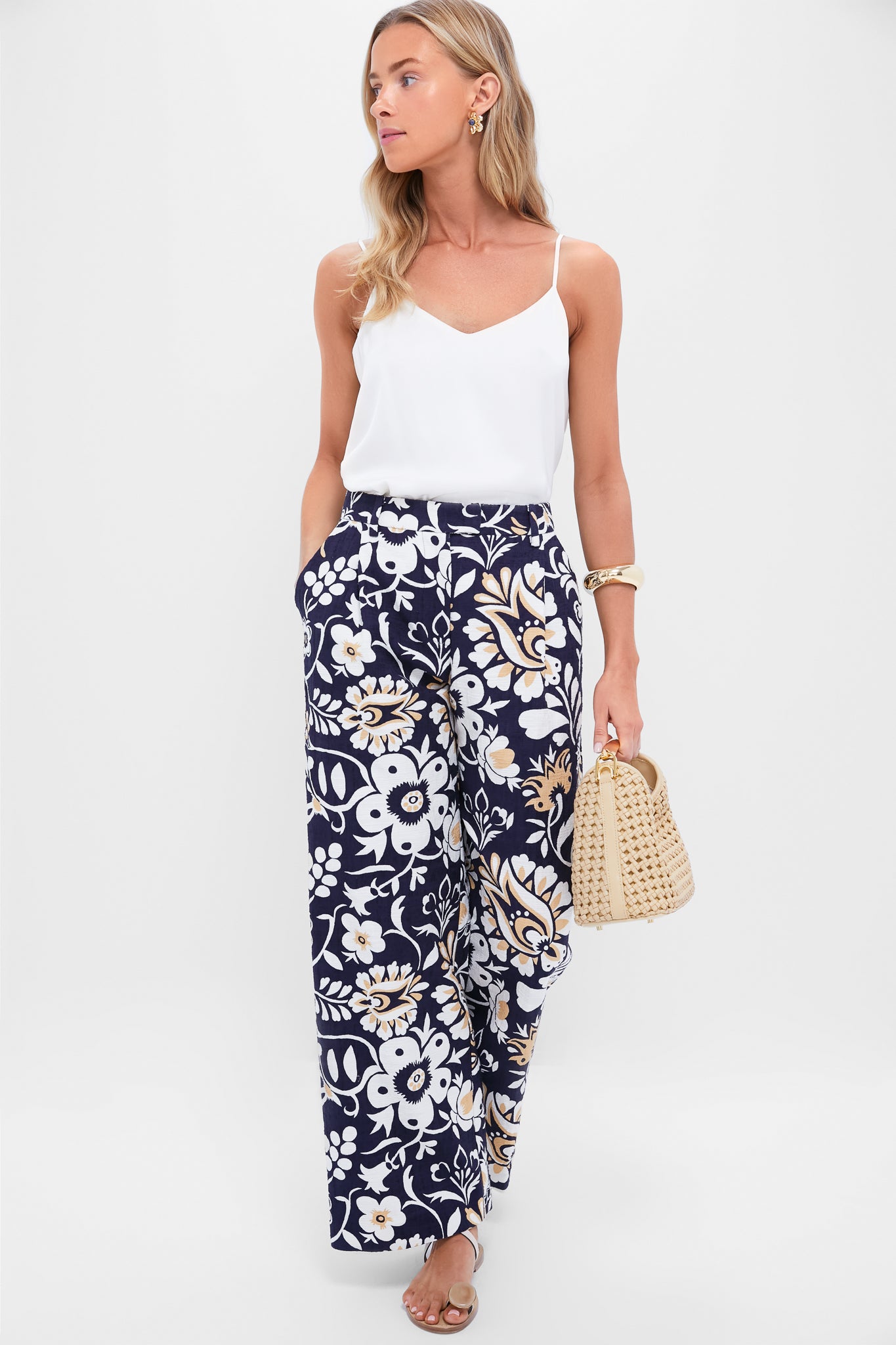 A woman showcases classic elegance with Gold Multi Lelani Stud Earrings, effortlessly pairing them with a chic floral outfit and woven straw handbag for a high-end, fun look.