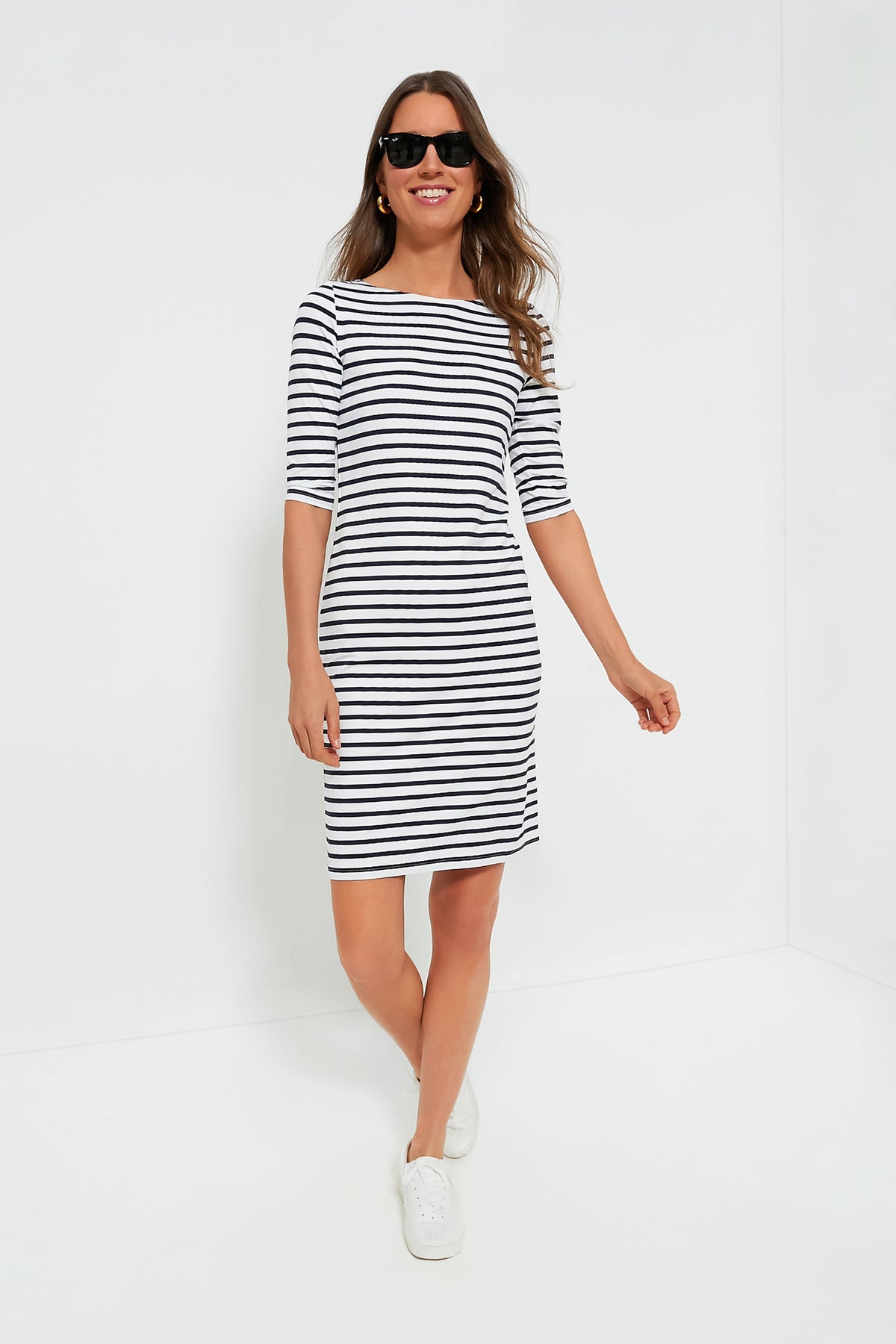 A woman in sunglasses and a Saint James black-and-white striped dress with elbow-length sleeves and white sneakers stands confidently, showcasing classic style.