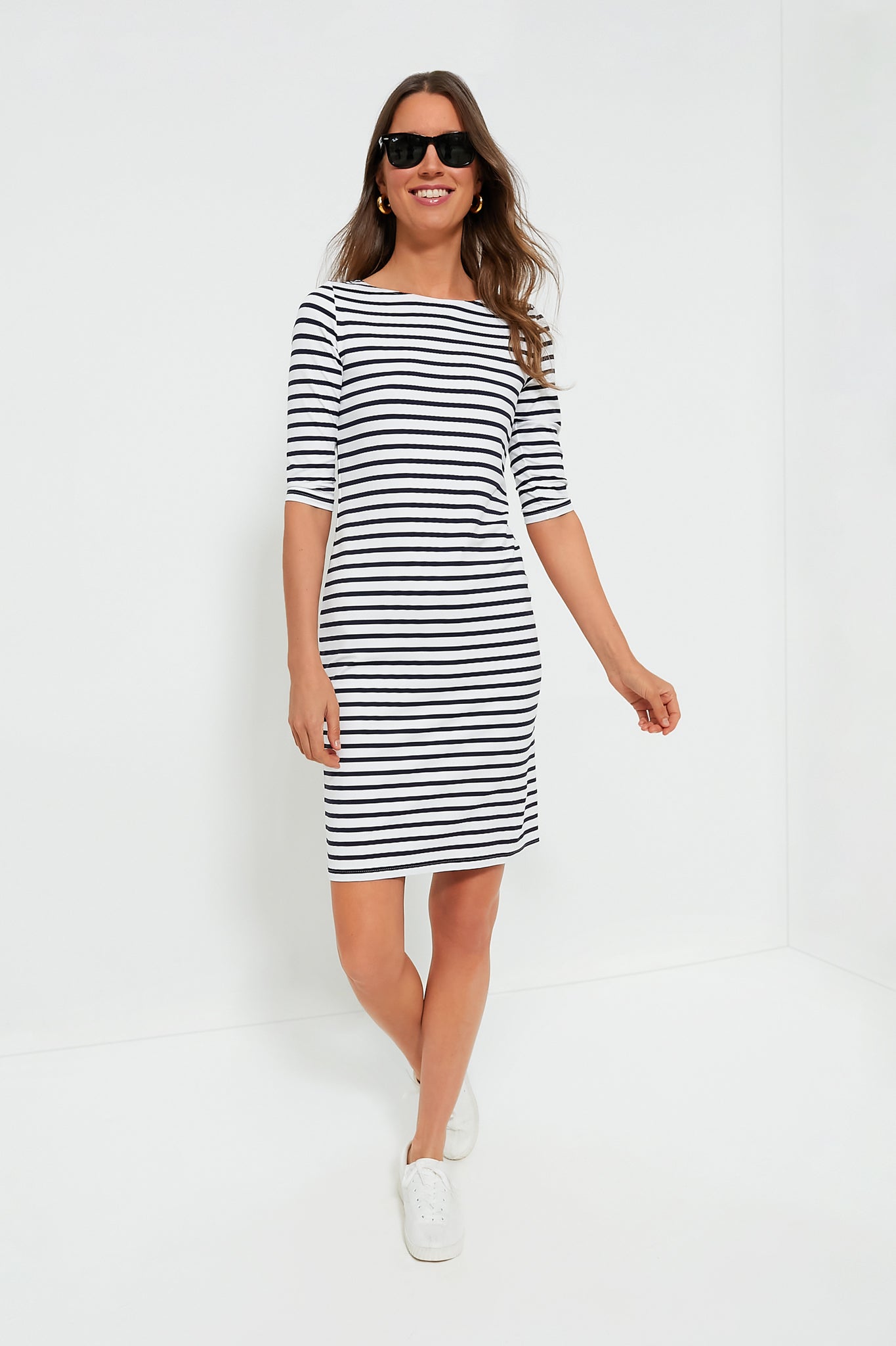 A woman in sunglasses and a Saint James black-and-white striped dress with elbow-length sleeves and white sneakers stands confidently, showcasing classic style.