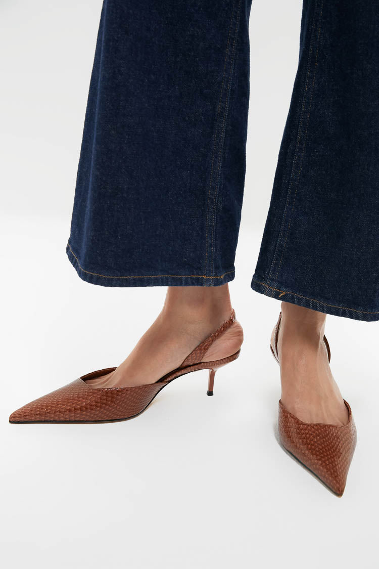 A person stands on a white surface wearing dark denim pants and Schutz brown textured slingback pointed-toe kitten heels.