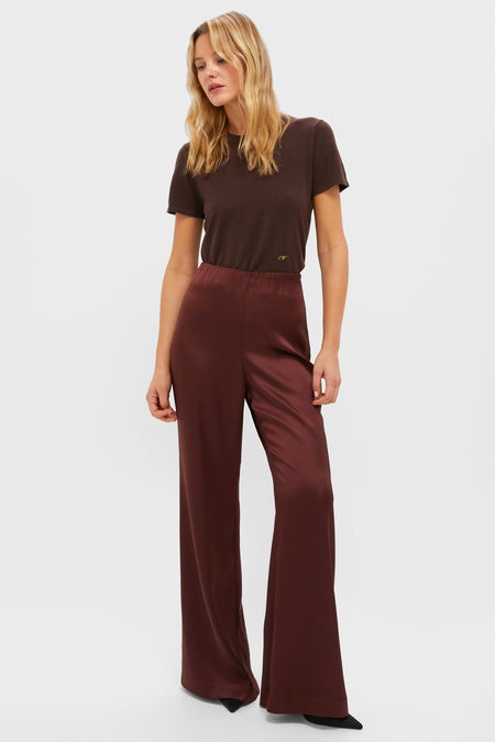A woman stands against a white background wearing a short-sleeved brown Tuckernuck t-shirt paired with high-rise, wide-leg maroon satin trousers.