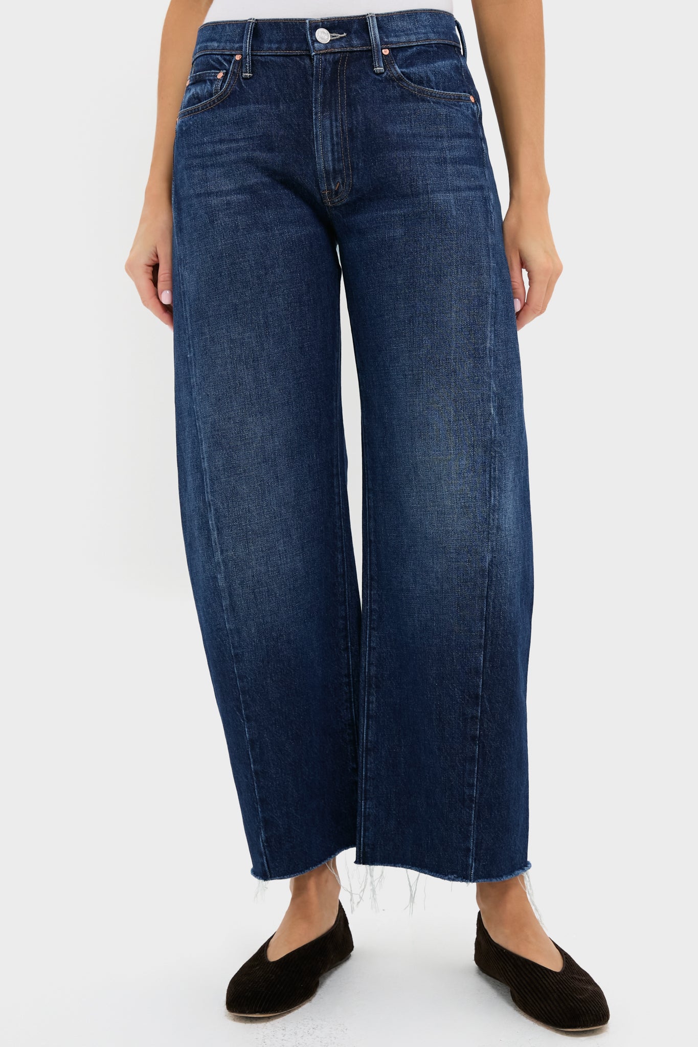 A person wears MOTHER high-waisted, dark blue barrel leg jeans with a frayed hem and black flats, standing in a simple white space.